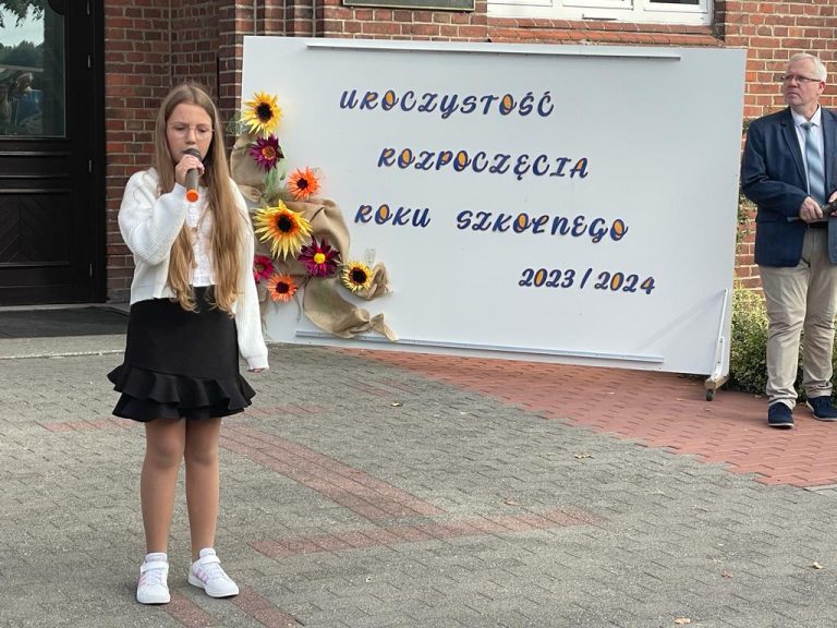 Uczennica klasy 5 a Joanna Kurosińska na tle szkoły i tablicy przedstawiającej napis uroczystość rozpoczęcia roku szkolnego 2023/2024