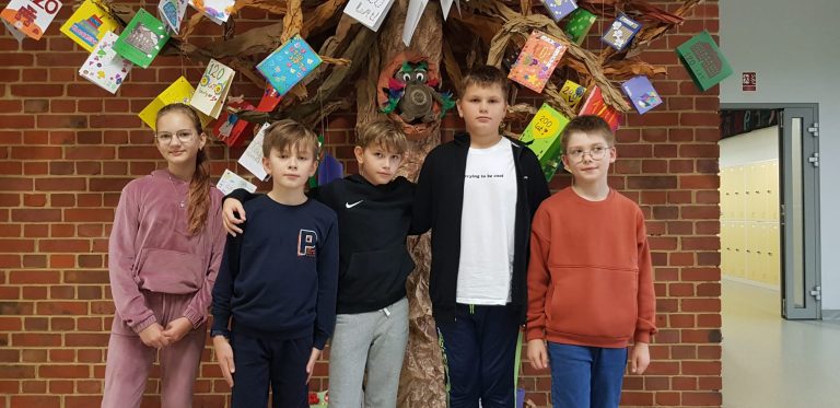 pięcioro uczniów klas czwartych na tle sztucznego drzewa zrobionego z papieru, na którym wiszą kartki przysłane do szkoły z okazji 120-lecia szkoły
